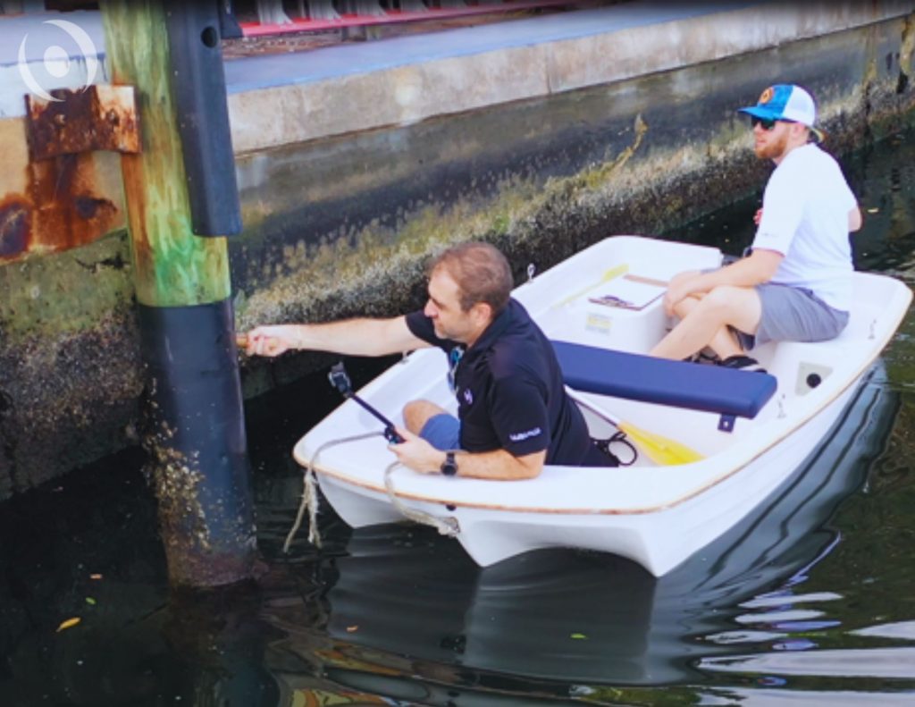 Two engineers inspecting seawall pilings from a small boat, evaluating corrosion and marine growth during a professional seawall inspection in South Florida.