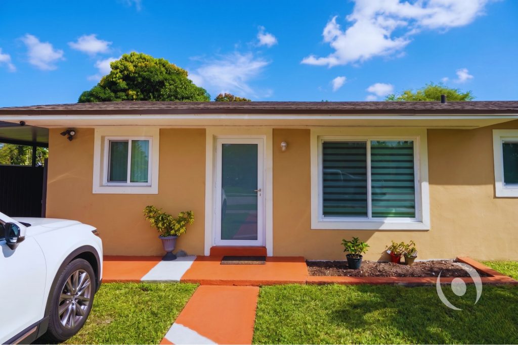 Front entry of completed home addition in South Florida showing new exterior door, impact windows, security lighting, and finished stucco facade engineered and designed by NOHMIS