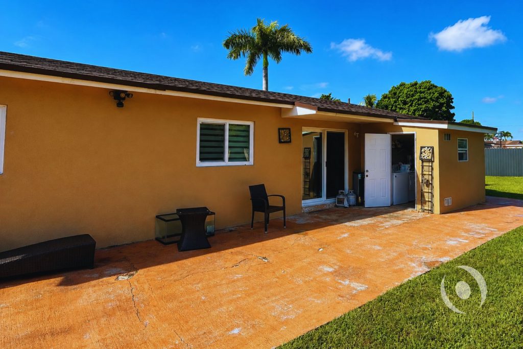 Completed home addition rear exterior in South Florida showing sliding glass door, exterior entry door, security flood lighting, and stucco finish designed and engineered by NOHMIS