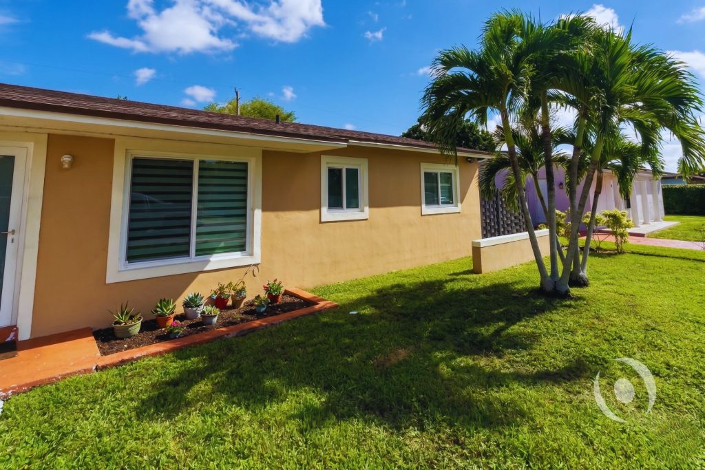 Side exterior of completed residential addition in Miami Gardens showing finished stucco walls, new impact windows, palm trees, and fresh landscaping designed by NOHMIS