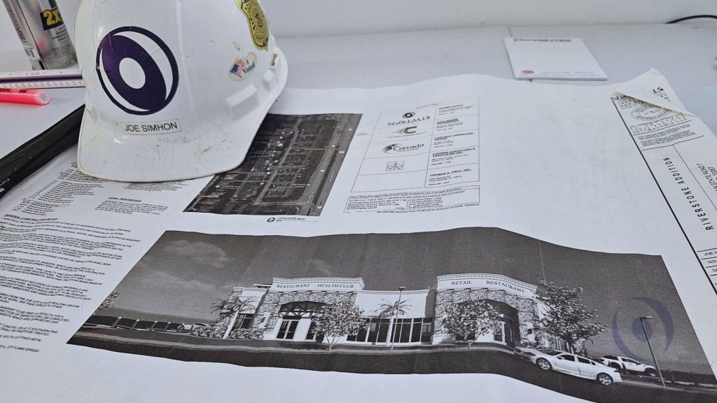 NOHMIS founder Joe Simhon's hard hat resting on architectural and structural engineering blueprints for the Riverstone Shoppes commercial building addition in Parkland, Florida