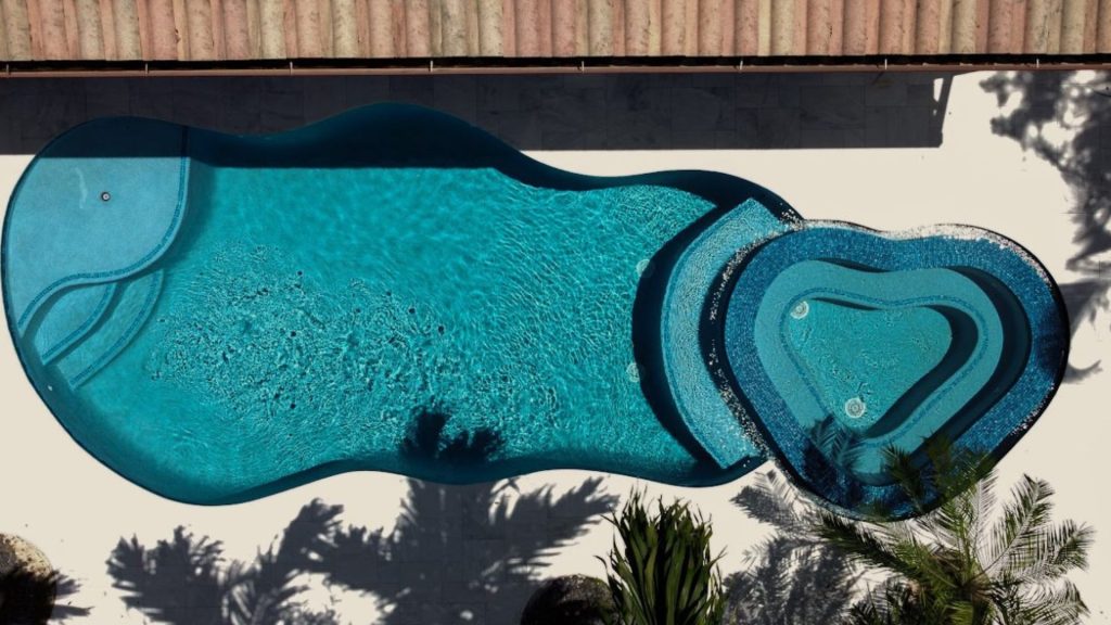 Aerial view of a custom freeform swimming pool with a raised spa, blue mosaic tile, and surrounding patio by Master Touch Outdoor Living.