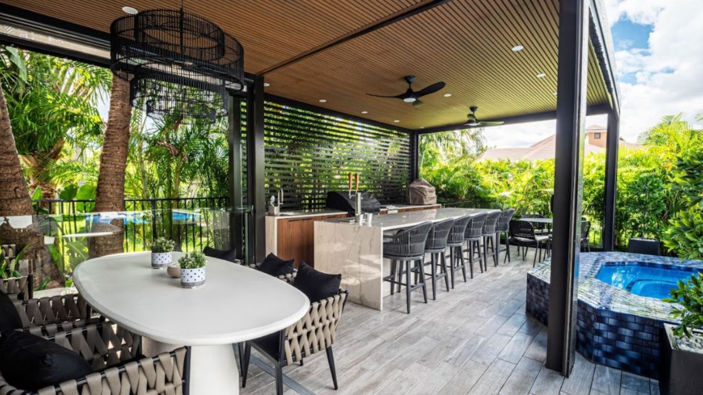 Custom covered outdoor living space with a wood slat ceiling, outdoor kitchen and bar, dining area, and tiled spa by Master Touch Outdoor Living.